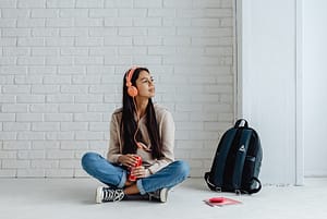 A young woman sitting on the ground, listening to music and next to a backpack